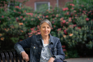Retiring professor Andrea Marks sits on a bench in front of rhododendrons